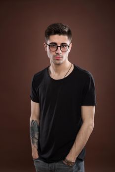 Young man in depression. Stylish male student wears round spectacles, has trendy hairstyle, looks confidently, isolated over brown background. People and human expressions