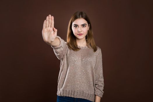Close up portrait of a serious young woman showing stop gesture with hand isolated over brown background