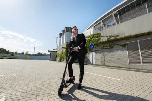 Man in a black business suit stands next to an electric scooter and talks on the phone.