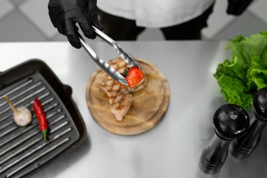 Close-up of the chef's hand as he puts a tomato from the grill on a plate with tongs.