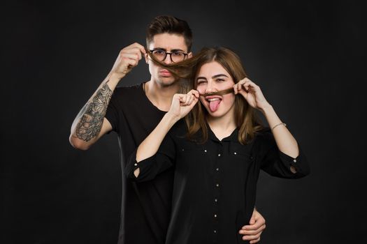 Beautiful young loving couple making fake moustache from hair while standing against black background. Funny moustache.