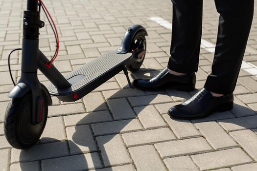 Close-up of a man's feet in a business suit and shoes near an electric scooter.