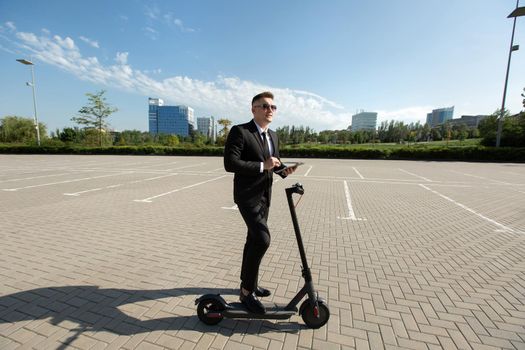 Young handsome businessman in a suit rides an electric scooter and looks at a laptop.