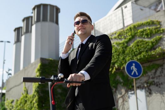 Man in a black business suit stands next to an electric scooter and talks on the phone.