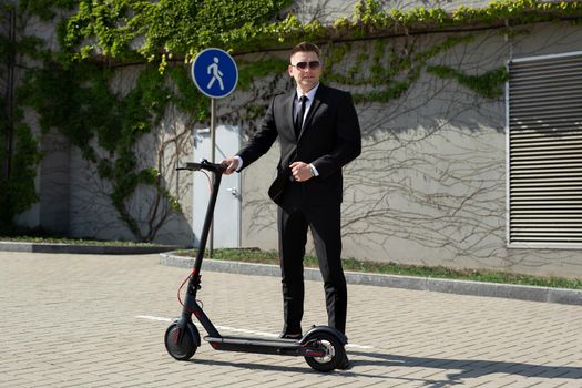 Man in a black business suit stands next to an electric scooter.