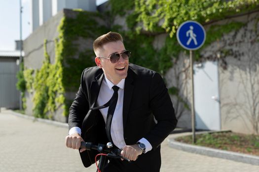 Man in a business suit and sunglasses rides an electric scooter and laughs.