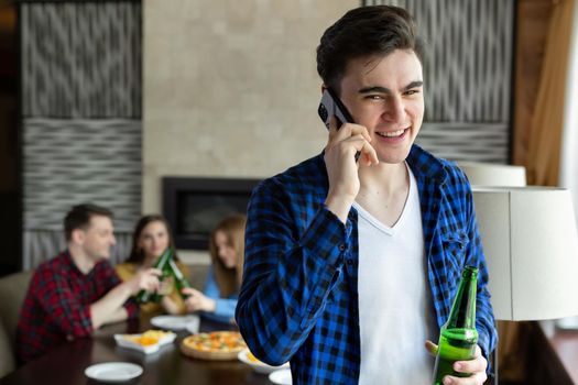 Young man drinks beer from a bottle, talks on the phone and looks out the window of a cafe against the background of his friends who are sitting at the table and eating pizza.