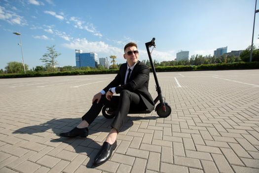 Young, handsome businessman in a suit sits on an electric scooter and laughs.
