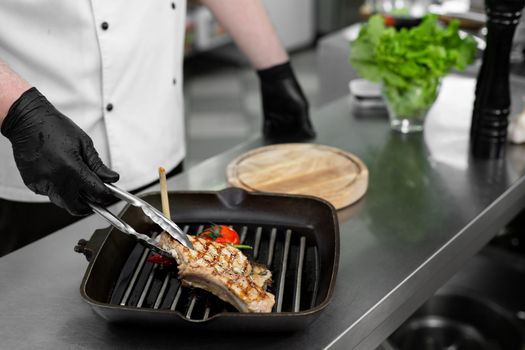 Close-up of a chef's hand holding a grill pan with grilled steaks and vegetables.