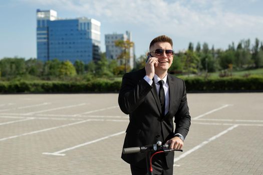 Man in a black business suit stands next to an electric scooter and talks on the phone.