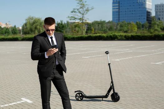 Young handsome businessman in a suit rides an electric scooter and looks at the phone.