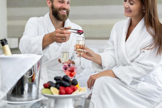 Man puts berries in a glass of sparkling wine to his woman in a hotel in bed.