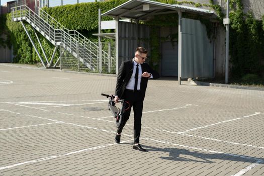 Young businessman in a suit looks at his watch and carries an electric scooter in his hands.