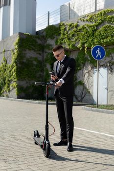 Man in a black business suit stands next to an electric scooter and holds a phone.