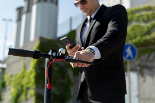 Man in a black business suit stands next to an electric scooter and holds a phone.