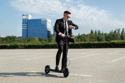 Young businessman with electric scooter standing in front of modern business building looking at watch.