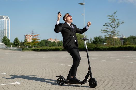 Young handsome businessman in a suit rides an electric scooter and is happy after a conversation on the phone.