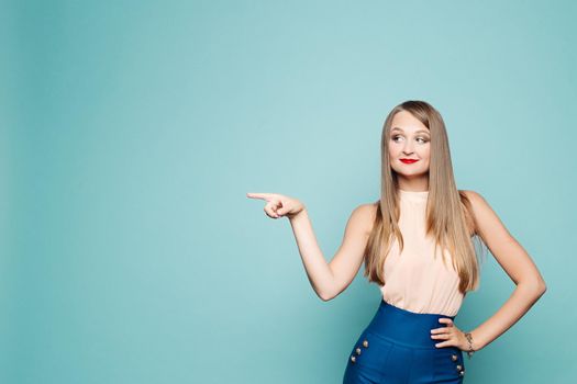 Studio portrait of blonde woman in top and elegant trousers pointing at something over blue background. Copyspace.