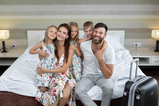 Young family: parents with children sit on the bed in the hotel and laugh. Traveling with children.