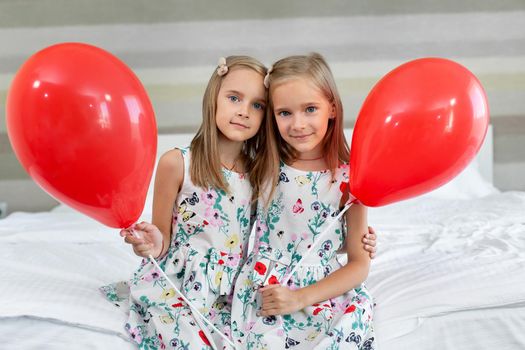 Two twin sisters in beautiful dresses are sitting on a bed in a hotel with balloons in their hands.
