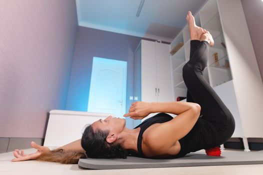 A middle-aged woman doing yoga makes herself a lower back massage with a massage ball while lying on the floor. Myofascial release.