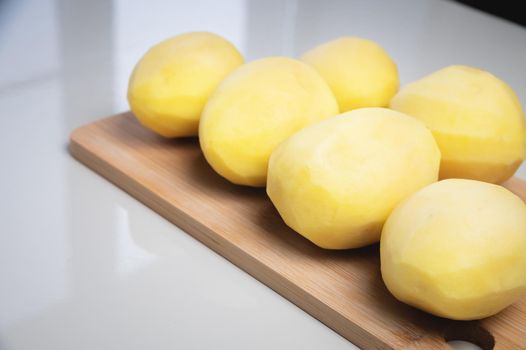 Peeled fresh potatoes on a wooden cutting board on a white table