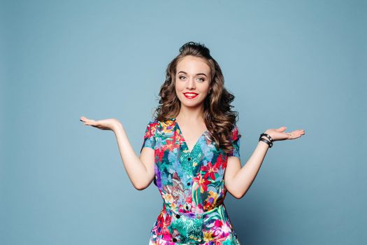 Portrait of lovely brunette with wavy hair in gorgeous summer dress demonstrating two objects over blue background.