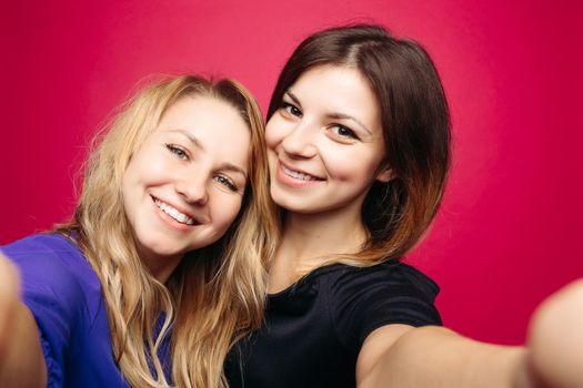 Self portrait of two beautiful, positivity girls, taking photo,smiling and posing together. Happy emotionally women with hairstyles,makeup, using camera at studio. Woman s friendship. Pink background.