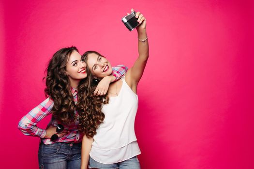 Two beautiful and fashionable women wearing in stylish clothes spending time together, making photo at camera. Positivity girlfriends posing, smiling and taking self portrait. Pink background.