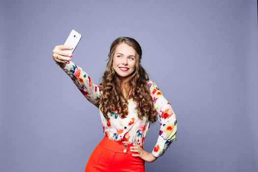 Studio portrait of beautiful trendy girl with long curly hair and make up holding arm on waist and taking selfie via cell phone over violet background. Cutout.