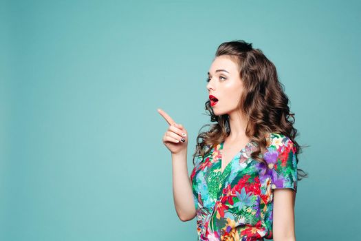 Portrait of shocked emotional curly brunette girl pointing by finger at side with opened mouth. Beautiful woman in floral dress surprised looking at side showing by hand. Concept of shopping.