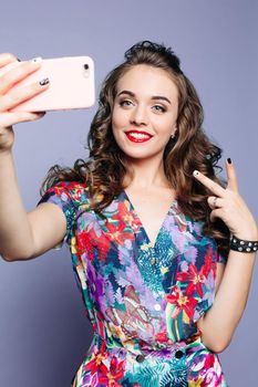 Fashionable woman in floral print dress smiling and taking photo at camera. Brunette girl with red holding call phone at hands and making self portrait of herself and showing peace. Green background.