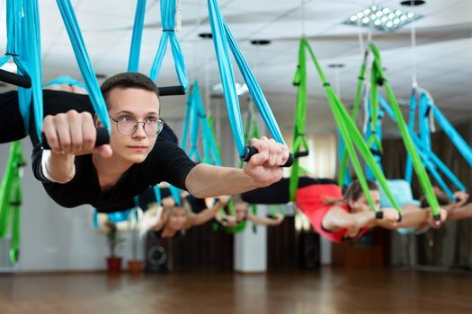 Group of young men and women do aerial yoga in hammocks at a fitness club.