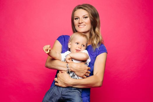 Studio portrait of loving mother with blonde hair embracing her playful baby over magenta background. Smiling at camera while baby moving in her hands.