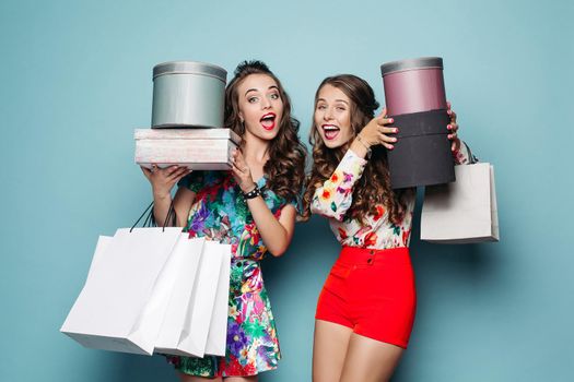 Studio portrait of two girlfriends with shopping bags after shopping. Two friends in trendy looks with wavy hair holding new clothes and hat in shopping bags over blue background.