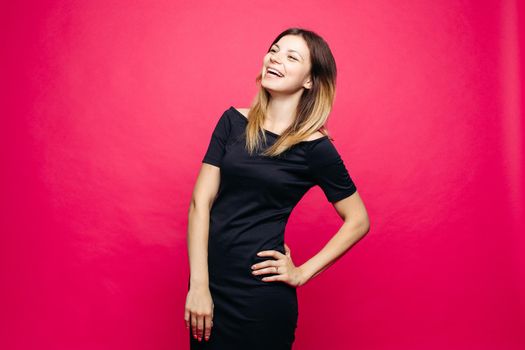 Happy young woman in black dress posing at studio and lauthing, looking away. Positivity, emotionaly, girl holding hand on waist, standing against pink background. Concept of shopping, fashion,beauty.