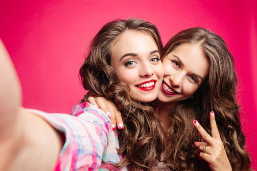 Self portrait of beautiful brunette girls holding camera in hand and tacking photo of herself. Young female with make up, in fashionable clothes making photo together. Pink studio.