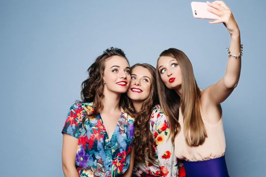 Studio portrait of smiling beautiful girlfriends wearing trendy outlooks with hairstyles and make-up taking self-portrait via cell phone over blue background.