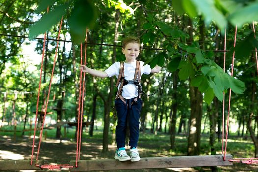 Small boy in climbing gear is walking along a rope road in an adventure Park, holding on to a rope and a carabiner.