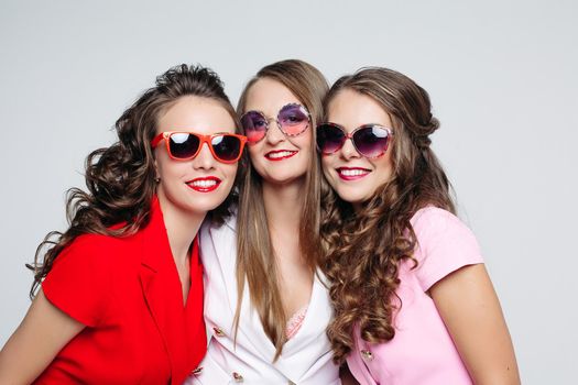 Studio portrait of beautiful long-haired girls in bright jackets and fashionable sunglasses smiling at camera against white background.