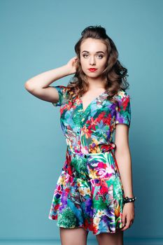 Studio portrait of beautiful young girl with red lips and wavy hair wearing lovely floral dress with hand at hair looking at camera over azure background.