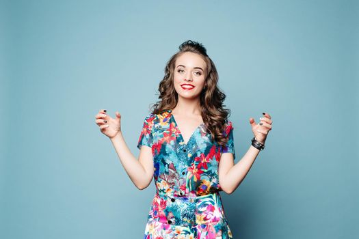 Studio portrait of attractive woman with long wavy hair and make up wearing trendy summer dress with flowers holding two invisible bottles in hands, smiling at camera. Isolate on blue background.