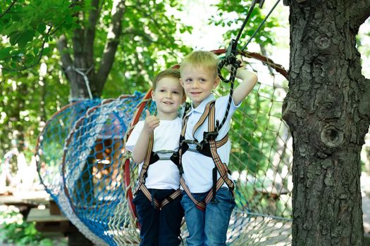 Two cute boys in an adventure park are doing rock climbing or passing obstacles on a rope road, hugging and giving a thumbs up.