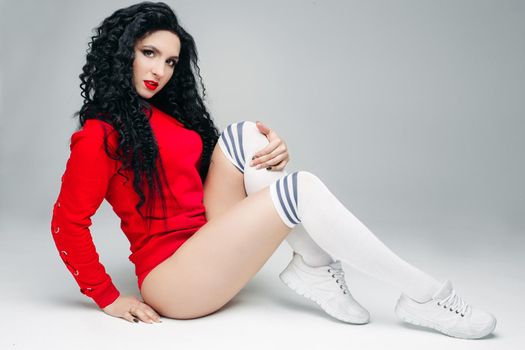 Portrat of sexy and attractive fit young brunette in red, sitting and looking at camera. Swag stylish model with curls long hair and red lips posing and touching leg. Concept of lifestyle sport.