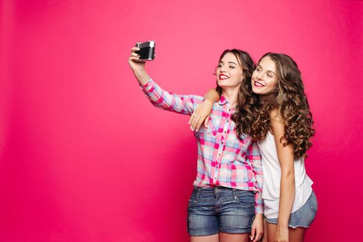 Studio portrait of beautiful ladies taking friendly selfie via vintage film camera over bright background. Hugging and smiling. Isolate.