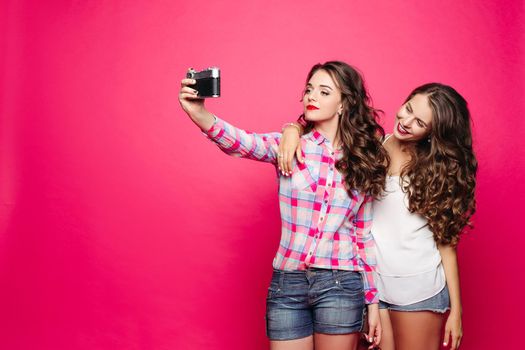 Studio portrait of beautiful ladies taking friendly selfie via vintage film camera over bright background. Hugging and smiling. Isolate.