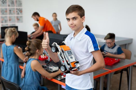 Portrait of a happy schoolboy boy with a robot that he himself assembled in robotics lessons at school, the boy shows a thumb up