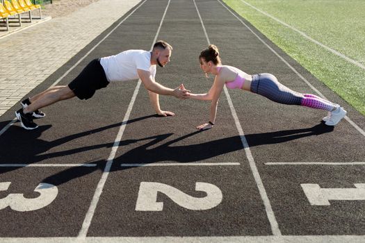 Sports active man and woman train in the summer outdoors in a public stadium, they stand in the plank and slap each other's hands.