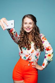 Positivity beautiful girl holding smart phone against face and tacking photo herself. Young female in fashionable wear, smiling, posing at camera and making self portrait. Blue studio background.