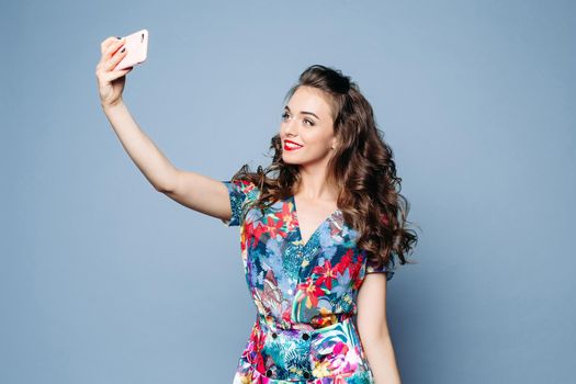 Studio portrait of beautiful smiling woman in floral dress with red lips and wavy hair making selfie over light blue background with cell phone.